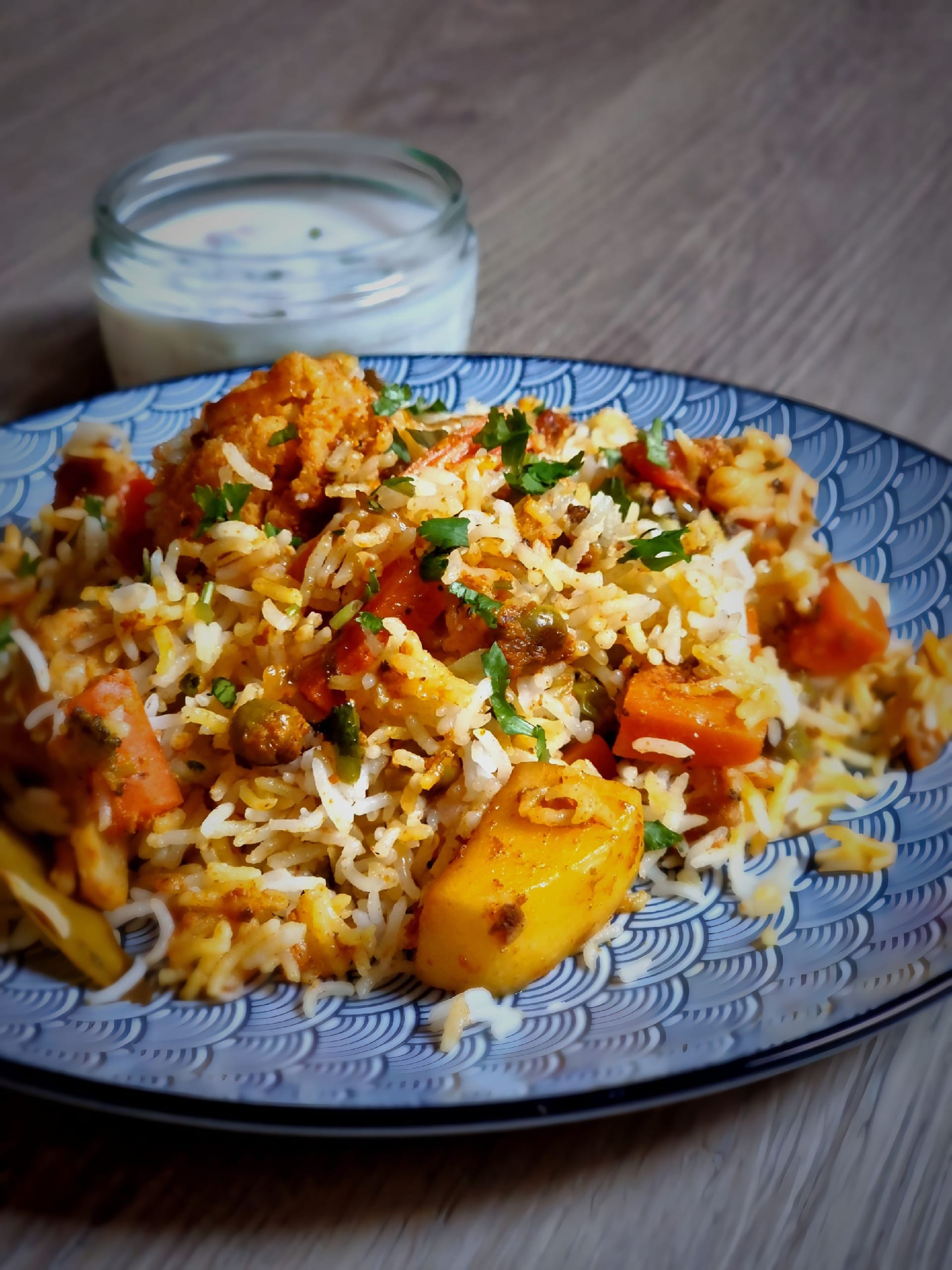 Vegetable biryani on a blue patterned plate — long-grain rice with carrot, potato, peas and cauliflower, scattered with coriander, a jar of raita behind it.