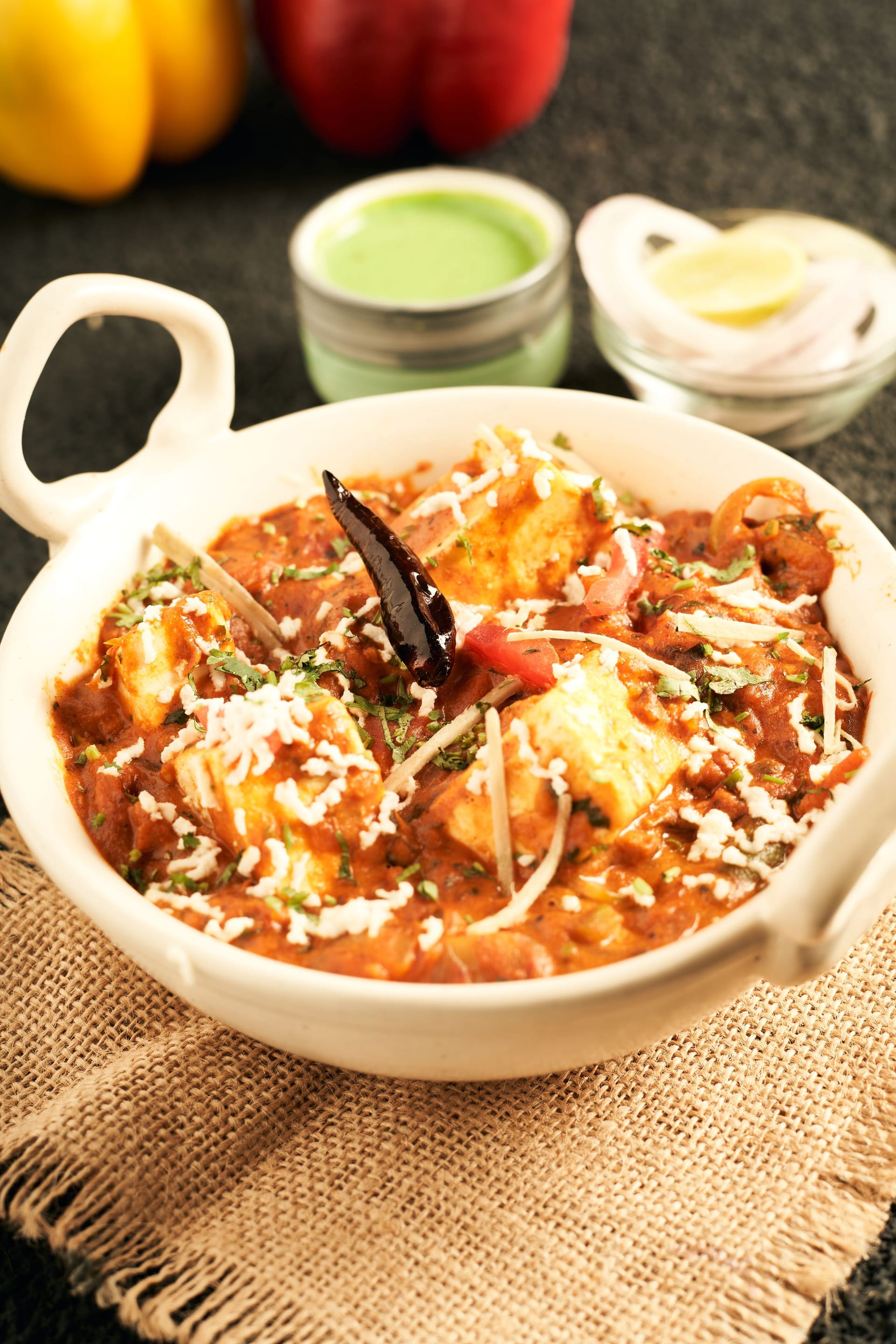 Paneer butter masala in a white handi — cubes of paneer in a tomato gravy under grated paneer, ginger juliennes and a dried red chilli, with mint chutney and onion salad behind.